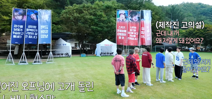 Stray Kids members, dressed in red and blue, stand in a field in front of banners for their teams. The red team has blue banners, and the blue team has red banners.