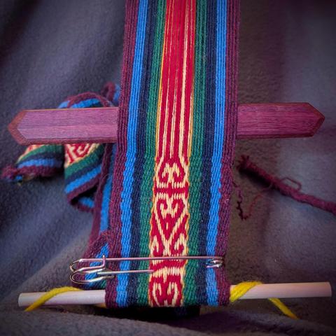 Woven band shows outside borders in solid stripes (burgundy, light blue, dark blue, and dark green) and inner pattern in red and yellow, with reflected and mirrored and flipped curled shapes. There is a weaving stick holding open the next row for the weft thread. The base of the weaving is safety pinned on a small dowel which is looped into a yarn belt.