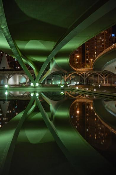 The image shows a nighttime scene underneath a bridge in Valencia’s City of Arts and Sciences. The modern concrete structure is dramatically lit by green lights. The curved beams and columns create a geometric pattern that is symmetrically reflected in the still water below. In the background, illuminated buildings of the city are visible. The composition appears architecturally complex and futuristic, with strong contrasts between light and shadow.