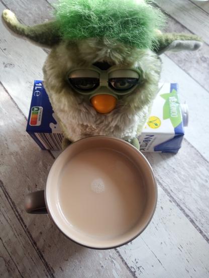 Das Bild zeigt einen Plüsch-Furby mit grünem Fell und großen Augen. Der Furby sitzt auf einem Holztisch und hat einen Becher mit einem beige gefärbten Getränk, das Kaffee mit Hafermilch ist, vor sich. Unter dem Furby liegt ein Hafermilchkarton. Der Tisch hat eine verwitterte, weiß gestrichene Holzoberflächenimitationstischdecke. Der Furby hat einen gelben Schnabel und wirkt etwas müde oder träge.