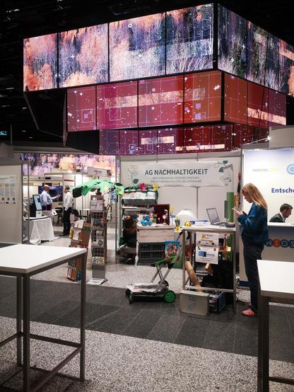 Stand der AG Nachhaltigkeit auf dem Bibliothekskongress Wien, rechts vorn eine blonde Fraue, die auf ihr Handy schaut, dahinter der Stand mit einem gebastelten Baum an dem Ideen stehen
