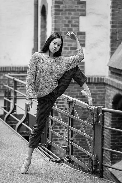 Eine Ballerina in normaler Straßenkleidung mit Spitzenschuhen steht auf einem Bein auf Spitze und hat das andere Bein auf ein Geländer in einem Innhof gestellt.