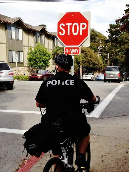 A police officer on a bicycle is parked on the side of the street at a stop sign. He is watching for offenders of the 4-way stop at this intersection in Carmel-by-the-Sea, California.