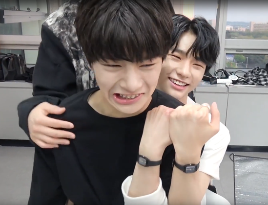 I.N sits on Hyunjin's lap. Felix's hand is visible on I.N's shoulder. Hyunjin and I.N are each holding up a wrist to show that they are wearing matching watches. Hyunjin looks happy and I.N is grimacing.