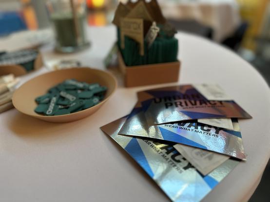 Das Bild zeigt eine Tischansicht mit verschiedenen Gegenständen. Im Vordergrund liegen mehrere glänzende Karten mit der Aufschrift "URBAN PRIVACY" und dem Slogan "WEAR WHAT MATTERS". Diese Karten sind in silber- und blau-gefärbten Farbtönen gehalten. Neben den Karten befindet sich ein beige Schalen, gefüllt mit grünen Einkaufswagenchips, die die Aufschrift "Ossrox" tragen. Im Hintergrund ist ein kleiner, brauner Karton mit einem Hausmodell zu sehen, in dem weitere grüne Einkaufswagenchips aufgereiht sind. Der Tisch ist weiß und die Umgebung wirkt wie ein Veranstaltungsort, möglicherweise ein Konferenzraum oder eine Ausstellung.
