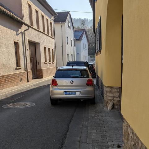 Silberner Volkswagen auf schmalem Kopfsteinpflaster-Gehweg neben gelbem Gebäude in ruhiger Dorfstraße im Morgenlicht auf Gehweg geparkt. Dahinter noch mehr solcher Falschparkenden