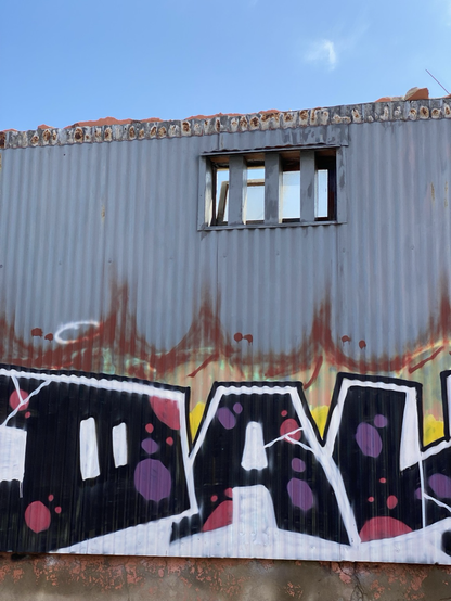 Mostly torn down building, only a Graffiti painted wall sheathed in corrugated metal with one small open rectangular window seen through bars.