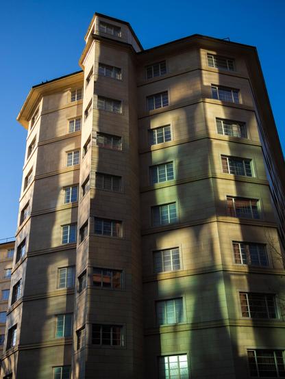 Photo looking up at an angular honey-coloured stone building, at least eight storeys high, on a sunny day. In the  middle of the wall a narrow, sharp extension sticks out, perhaps around a staircase, with, on every floor, windows either side of the point. There are also windows on either side as well as on the wall's chamfered edges. The whole face of the building, which would otherwise be shadowed towards the right, is dappled with green and white bands of light reflected from the windows of another building, probably behind the camera. Around the top is a bright blue sky.