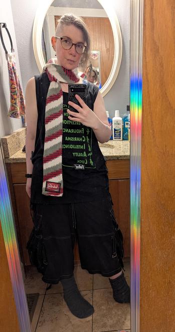 Image depicts a person shown in a full length bathroom door mirror, holding a spiderweb print cased phone taking a pic. Person has an undershaved  hairstyle in a ponytail, is wearing glasses, and is wearing a black tank top and shorts. A red, black, grey, and white diagonally striped knitted scarf is wrapped around their neck. The patch at the bottom of the scarf is similarly colored and reads "Nuka-Cola" in a flowing script font.