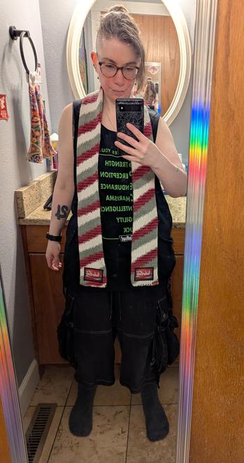 Image depicts a person shown in a full length bathroom door mirror, holding a spiderweb print cased phone taking a pic. Person has an undershaved  hairstyle in a ponytail, is wearing glasses, and is wearing a black tank top and shorts. A red, black, grey, and white diagonally striped knitted scarf is wrapped around their neck. The patch at the bottom of the scarf is similarly colored and reads "Nuka-Cola" in a flowing script font.