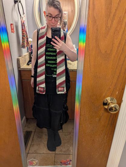 Image depicts a person shown in a full length bathroom door mirror, holding a spiderweb print cased phone taking a pic. Person has an undershaved  hairstyle in a ponytail, is wearing glasses, and is wearing a black tank top and shorts. A red, black, grey, and white diagonally striped knitted scarf is wrapped around their neck. The patch at the bottom of the scarf is similarly colored and reads "Nuka-Cola" in a flowing script font.