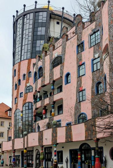 A medium shot captures the whimsical and colourful facade of the Hundertwasserhaus (The Green Citadel) in Magdeburg, Germany. The building boasts an irregular and organic design, characteristic of Friedensreich Hundertwasser's architectural style. The primary colours are pink and cream, with vertical stripes of darker, rough-textured material. Windows of various shapes and sizes, many framed with bright blue, are scattered across the facade seemingly at random.
The building features multiple levels, with some balconies adorned with colourful, bulbous elements and greenery. A prominent cylindrical tower with numerous dark-framed windows rises from the left side of the structure, contrasting with the more pastel tones of the main building. The roofline is uneven and features decorative elements.
At ground level, there are arched doorways and colourful, mosaic-like pillars supporting an overhang.