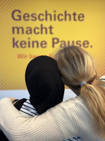 Bildeindrücke aus dem Projekt "Studio Cosmo": Eine Aufnahme von zwei Frauen, die sich vor einem gelben Poster umarmen. Sie sind von hinten aufgenommen und haben ihre Köpfe aneinander gelehnt. Auf dem Poster ist eine Aufschrift zu lesen: "Geschichte macht keine Pause."'