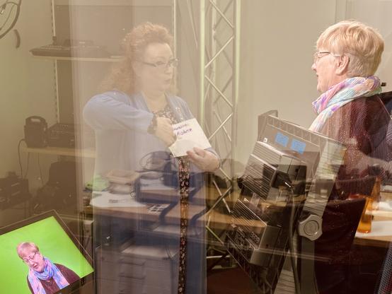 Zwei Frauen sprechen miteinander in einem Fernsehstudio. Eine Frau steht vor dem Greenscreen und spricht zur Kamera. Die andere Frau hält einen Zettel mit Text und scheint der Frau vor der Kamera Hinweise zur Aussprache zu geben.