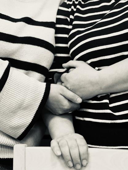 Eine Nahaufnahme in schwarz-weiß: Zu sehen sind die aneinander gelehnten Oberkörper zweier Frauen mit gestreiften Pullovern. Beide haben jeweils einen Arm vor dem Körper angewinkelt. Ihr Hände liegen sanft aufeinander.