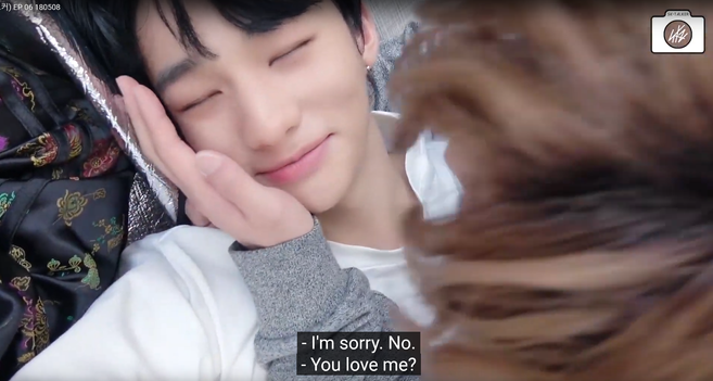 Hyunjin lies on his back with his eyes closed and a pleased smile. Lee Know is beside him and leaning over him, holding the back of Hyunjin's neck with one hand, and is gently holding the side of Hyunjin's face with his other hand. Caption: "I'm sorry. No." "You love me?"