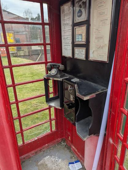 Interior view of an old British red public telephone kiosk with a Bakelite 200 type telephone and a “button A and B” pay phone mechanism. There are notices in frames on the wall above the telephone. Through the red glazing bars of the window an area of grass is seen with some brick buildings, a yellow advertising sign, and part of a red and white streetworks safety barrier.