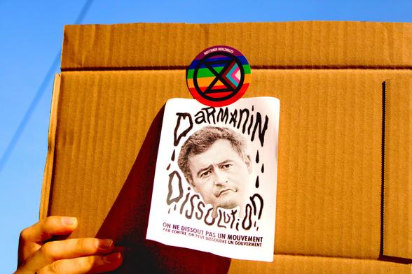 Sticker "Extinction Rébellion" avec le drapeau queer scotchant une affiche "Darmanin Dissolution" sur une pancarte