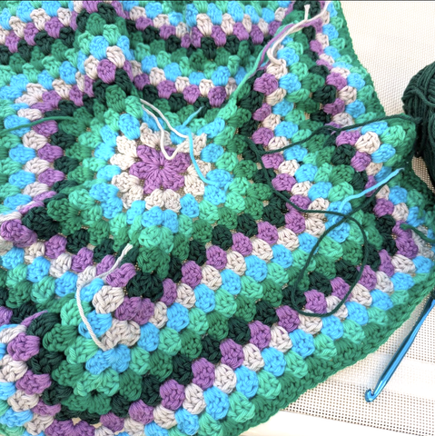Ein Häkelstück mit Hexagonalem Muster und in verschiedenen Pastelfarben gehalten. Lose Fäden hängen herum. Ein Wollknäuel und eine Häkelnadel sind zu sehen
