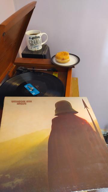 Record player with a 12" black vinyl record.
In the foreground is an album cover showing an ancient warrior figure, wearing a red robe and a greek helmet and carrying a spear, looking over hills bathed in the light of a low sun.
Behind the player is a mug of coffee on a speaker. On the corner of the player is a delicious jam doughnut.