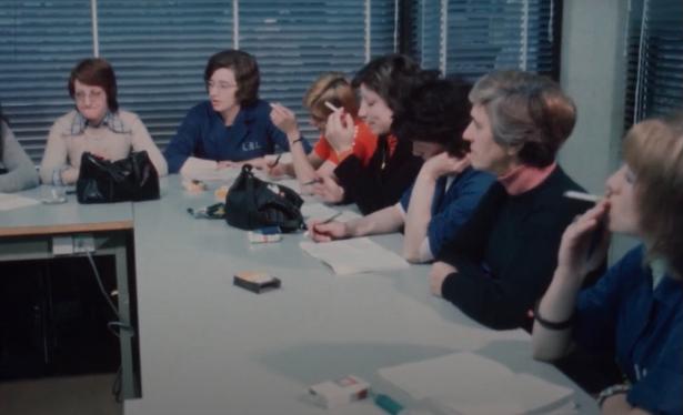 A still image from a documentary about the Liberated Broom cooperative. It shows a bunch of women sitting around a table in a meeting situation, possibly in the 70s or 80s, several of them smoking cigarettes and taking notes. Office setting, with closed blinds in the background.