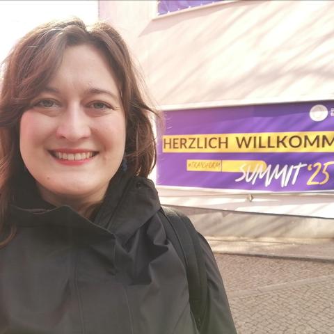 Laura mit langen braunen Haaren und breitem Grinsen vor einem Haus, an dem ein lila Banner hängt. Auf dem Banner steht "Herzlich willkommen! #transform_D Summit'25"