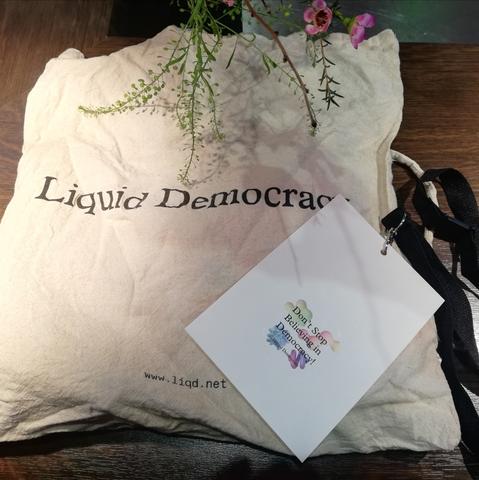 Heller Jutebeutel mit Aufdruck "Liquid Democracy" und "www.liqd.net". Darauf liegt ein kleines weißes Schild an einem schwarzen Schlüsselband. Auf dem Schild ist ein bunter Aufkleber. Auf dem steht "Don't Stop Believing in Democracy".