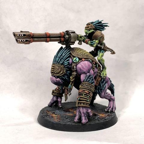 Large purple beast with a beak-like face wearing leather armor adorned with baubles. A green, smaller, thinner humanoid rides on top aiming a cannon mounted to the beast’s back. They stand on molten ground.
