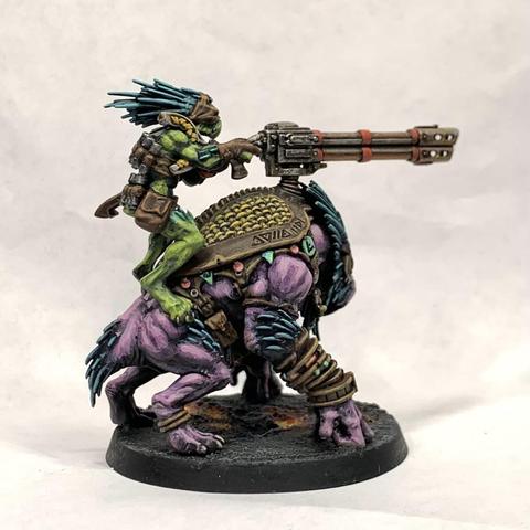 Large purple beast with a beak-like face wearing leather armor adorned with baubles. A green, smaller, thinner humanoid rides on top aiming a cannon mounted to the beast’s back. They stand on molten ground.