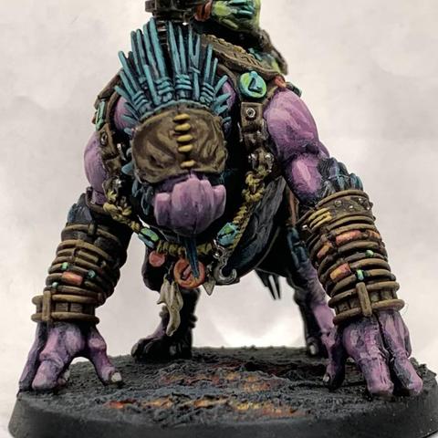 Large purple beast with a beak-like face wearing leather armor adorned with baubles. A green, smaller, thinner humanoid rides on top aiming a cannon mounted to the beast’s back. They stand on molten ground.