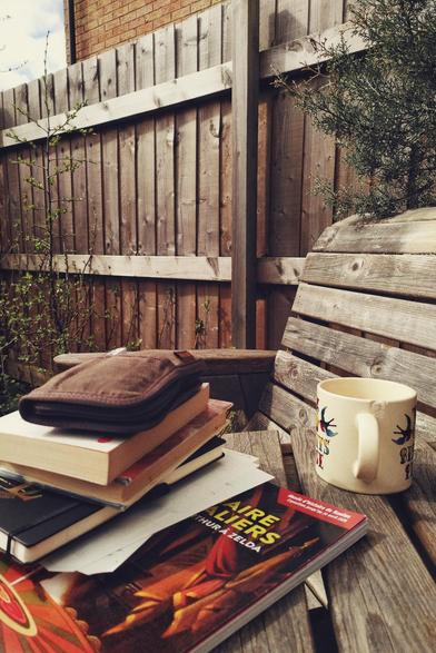 A vertical colour photograph of a small pile of books and notebooks on a wooden garden table. On top of it all is a pen pouch. Next to the pile is a mug seemingly empty. Behind is a fence and trees and bushes with young green leaves on.