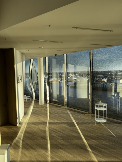 Der Blick geht aus den Fenstern der Elbphilharmonie auf den Hafen. Die Sonne scheint und es spiegeln sich die glänzenden Metallelemente auf dem Holzboden