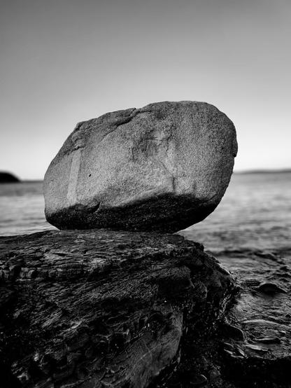 A black and white photo showing a large boulder perched on the rocky coast of Maine.