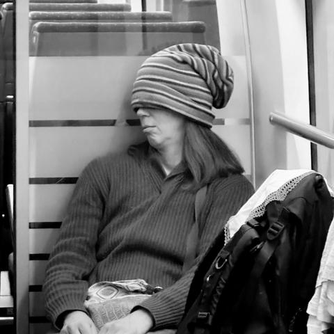 Eine Frau die ihre lange Mütze über die Augen gezogen hat und schläft. Sie sitzt in einem Zug. Das Foto ist in Schwarzweiß.