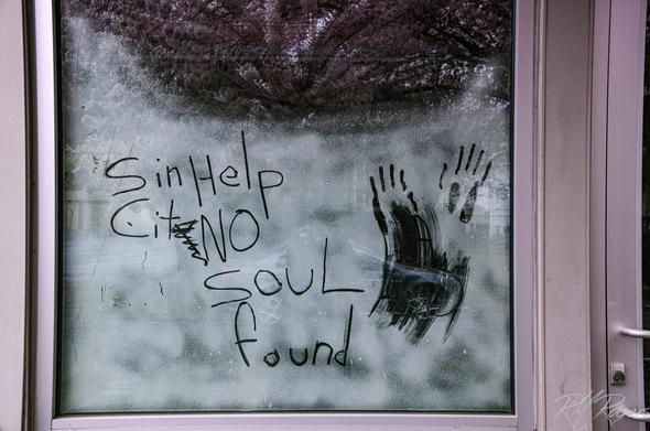 Photograph of a storefront window with the words “Send Help City No Soul Found” misspelled, scratched into the frosting, accompanied by fingernail drag marks.