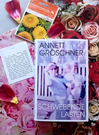 Foto des Buches, auf dessen Foto ein ausgeblichenes, verschwommenes Foto von zwei Frauen mit einem kleinen Kind im Arm zu sehen ist. Sie stehen vor einem weißen Lattenzaun. Die vordere, jüngere Frau trägt eine Sonnenbrille, ihre Haare werden von einem breiten weißen zurückgehalten. Sie hat eine Hand zum Kopf geführt und lacht in die Kamera. Das Buch liegt auf zwei verschiedenen Geschenkpapieren, die dicht an dicht mit Rosen bedruckt sind; links rosafarbene, die damit die Farbe des Covers aufnehmen, rechts welche in Rottönen. Oben links neben dem Buch liegt ein Saatguttütchen für Zwergsonnenblumen, darunter ein Zettel, auf dem folgendes Zitat aus dem Buch zu lesen ist: »Das von einer dichten Reihe Zwergsonnenblumen begrenzte Grundstück am Johannisberg sollte Hanna nicht mehr erleben, sie starb Anfang Juni, aber wann immer ihre Töchter in diesem Sommer zum Elbufer gingen, wunderten sie sich über die Sonnenblumen, und Barbara sagte, das ist doch das Grundstück, wo Muttis Laden war, und Selma sagte, seltsam, als hätte Gott ihr die Blumen geschickt. Aber Elisabeth wollte von Gott nichts wissen, auch wenn sie für die Blumen keine Erklärung hatte. ‚Vielleicht ein Kunstwerk.‘«