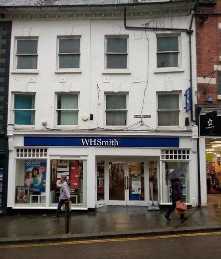 A white Victoria Shop front with a WH Smith shop on ground level and residential space above. The shop is on a slope and a couple of pedestrians are passing by.