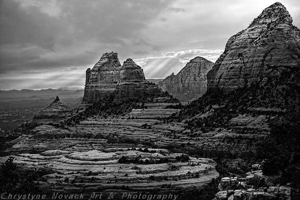 A dramatic black and white of the Sedona, Arizona landscape captures rugged, layered rock formations under a moody sky. Sun rays break through clouds, highlighting the texture and depth of the rocky terrain.