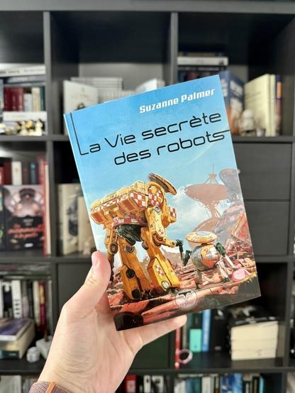 La couverture de La vie secrète des robots montre deux robots se tenant par ce qui leur sert de main. L'un est tout petit et porte un petit sac rose, le plus grand robot semble baisser le regard sur le petit. Le décor montre des vaisseaux et satellites.