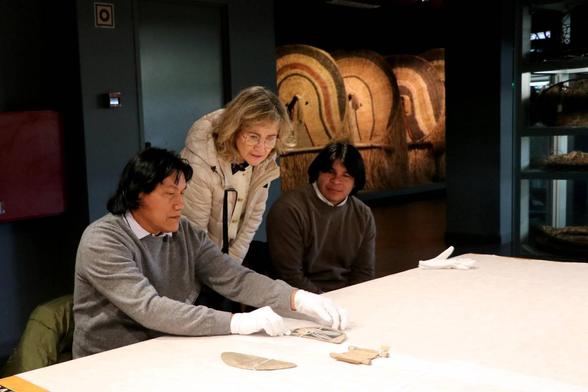 Kanawayuri Marcello Kamaiurá handling some pieces from the Kamayurá people on a table at the National Museum of Ethnology, with Elisabete Pereira and Auakamu Kamayurá at his side watching.
