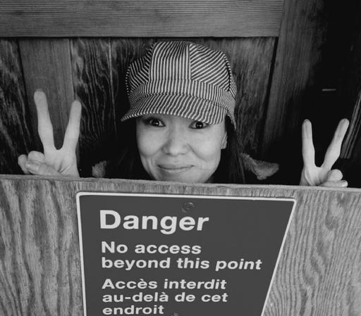 Portrait of me in a striped hat and smiling mischievously peers over a wooden barrier marked with a "Danger - No Access beyond this point" sign (translated in French). They are holding up both hands, making a peace sign with each. The photograph is in black and white, providing a timeless or documentary feel. The wooden structure framing the scene adds to the sense of containment and the juxtaposition of the playful figure with the warning sign creates a lighthearted, rebellious tone.