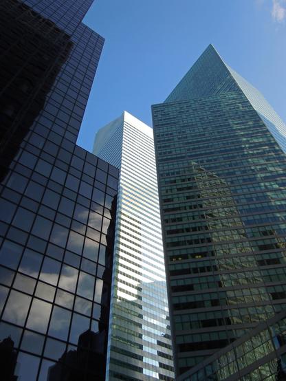 Color photo looking up at three tall office buildings, each one reflecting both the sky and other nearby towers