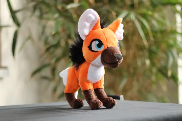 A little maned wolf plushie standing on grey table cloth. It's made of orange, white and brown short fur fabric and a long black mane.