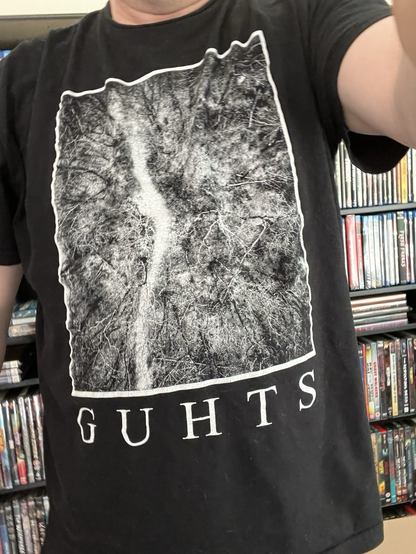 The image shows a person wearing a black T-shirt with a graphic design on the front. The design features a black and white aerial view of a forest with a prominent white river running through it, bordered by a white outline. Below the graphic, the word "GUHTS" is printed in white capital letters. The person is standing in front of a bookshelf filled with DVDs or video games, suggesting an indoor setting. The focus is on the T-shirt, with the background slightly blurred.