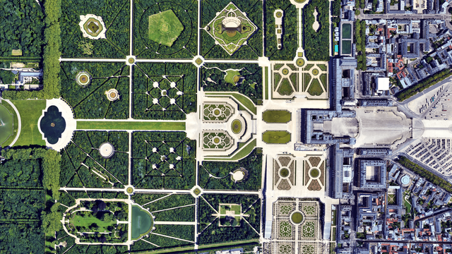 A zoomed in  satellite picture of the castle of Versaille and parts of its french garden. The castle is on the right side of the picture, with a grey blueish roof. The garden is very geometric with intricate green patterns, basins, trees and lawns.