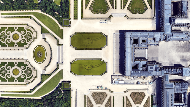 An even more zoomed in  satellite picture of the castle of Versaille and parts of its french garden. The castle is on the right side of the picture, with a grey blueish roof. The garden is very geometric with intricate green patterns, basins, trees and lawns.