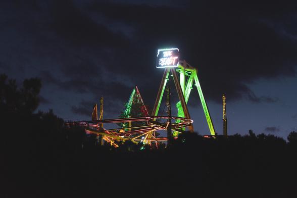 The image shows a nighttime scene featuring a brightly lit amusement park ride or roller coaster called "The Beast." The ride is illuminated with colorful lights, and a neon sign with the name is visible on top. The sky is dark with some clouds, creating a dramatic backdrop for the ride.