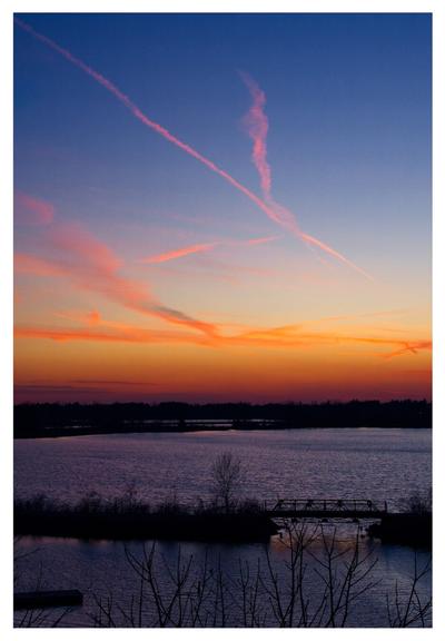 Pink and golden sunset over a dark lake. Trail streak across the deep blue sky.