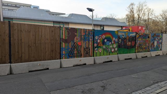 A wooden fence, street and pedestrian walkway. The wooden fence has a bunch of color full paintings.