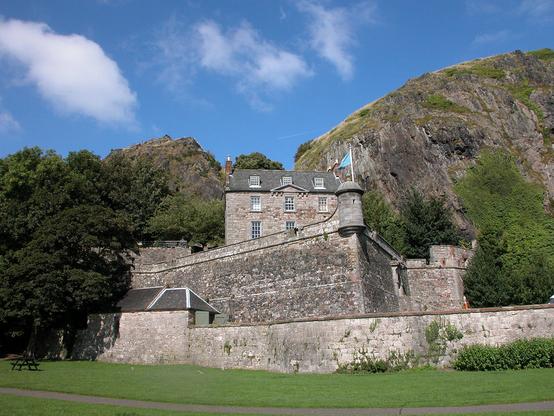 Dumbarton Castle. The image shows a grassy area in the foreground, beyond which is a low stone wall and then a higher stone wall with defences and a right angle facing the viewer. Above that is a stone house with a slate roof and attic windows. Behind that is a large cliff on the right and a smaller rock on the left. There are trees to the left of the walls. The sky is mainly blue.